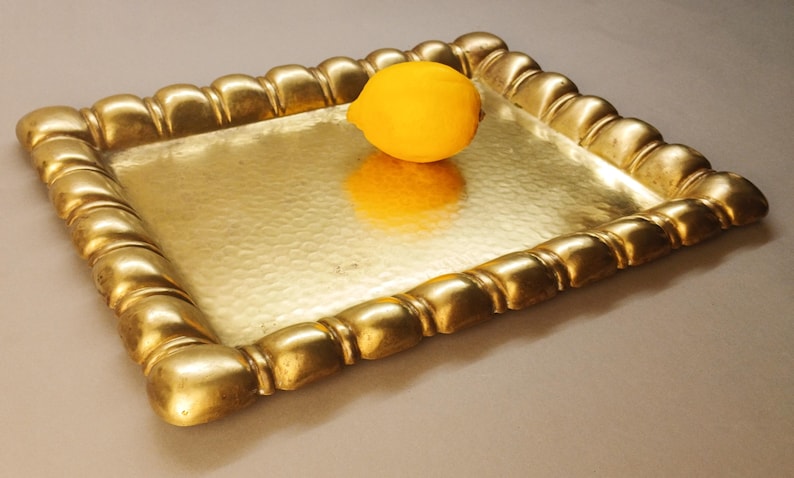 May include: A gold-colored metal tray with a hammered surface and a decorative, rounded border. A yellow lemon sits in the center of the tray, reflecting on the surface. The tray is rectangular and appears to be made of brass.