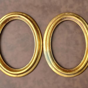 Könnte beinhalten: Zwei ovale, goldfarbene Bilderrahmen mit gerilltem Design und einem Used-Look-Finish. Die Rahmen werden vor einem braunen Hintergrund präsentiert. Jeder Rahmen ist etwa 15 cm lang.