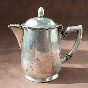 Op de afbeelding: Een vintage zilverkleurige koffiepot met een deksel en handvat. De pot heeft een tuit en een klein embleem op de voorkant. De pot is waarschijnlijk gemaakt van zilver of een zilverkleurig metaal. De pot staat op een bruine achtergrond.