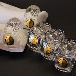 Op de afbeelding: Een set van vier servetringen van helder kristal met een waaiervormig ribbelpatroon. Elke ring heeft een ronde goudkleurige en zwarte sticker met de tekst Villeroy & Boch Crystal 24% PbO. De ringen liggen naast een wit servet met kanten rand.