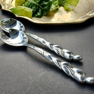 Puede incluir: Un par de brillantes cubiertos plateados para ensalada con un diseño trenzado en los mangos. Las cucharas para servir están junto a una bandeja dorada con lechuga verde. Los cubiertos están sobre una superficie oscura.