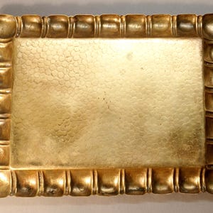 May include: A rectangular, antique-style tray with a textured surface and a decorative border. The tray is a warm gold color, with a hammered texture on the inside and a series of square-shaped accents around the edges.