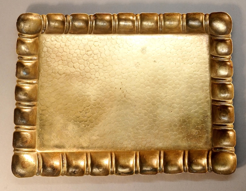 May include: A rectangular, antique-style tray with a textured surface and a decorative border. The tray is a warm gold color, with a hammered texture on the inside and a series of square-shaped accents around the edges.