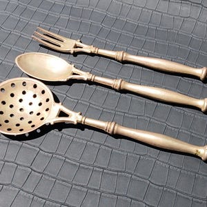 Puede incluir: Tres utensilios de servir antiguos de plata: un tenedor, una cuchara y una cuchara ranurada. Cada pieza tiene un mango decorativo y un diseño detallado. Los utensilios están dispuestos sobre una superficie oscura texturizada.