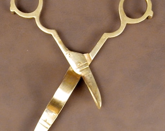 May include: A pair of gold-colored scissors with rounded handles and pointed blades. The scissors are made of a shiny metal and are set against a brown background. The blades are slightly open.