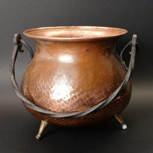 May include: A round, hammered copper cauldron with a wide, open rim. The vessel features a dark, curved metal swing handle attached to the sides and rests on three small, tapered metal feet. The copper surface has a polished, reflective finish.