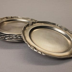 Puede incluir: Un conjunto de seis fuentes ovaladas plateadas con bordes festoneados y ornamentados. Las fuentes est&aacute;n apiladas sobre una superficie gris neutra, con una fuente ligeramente separada, mostrando los detalles del dise&ntilde;o.
