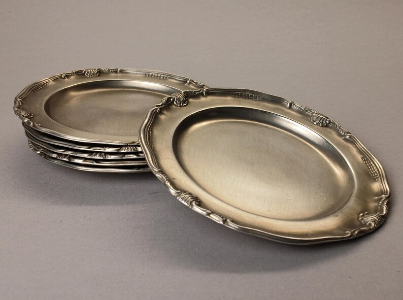 Puede incluir: Un conjunto de seis fuentes ovaladas plateadas con bordes festoneados y ornamentados. Las fuentes est&aacute;n apiladas sobre una superficie gris neutra, con una fuente ligeramente separada, mostrando los detalles del dise&ntilde;o.