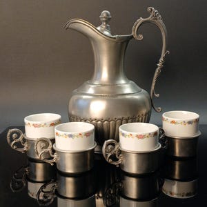 Op de afbeelding: Een zilverkleurige kan met een decoratieve handgreep en vier kopjes. De kopjes zijn wit met een bloemenpatroon en staan in zilveren houders. De kan en kopjes staan op een reflecterend oppervlak.