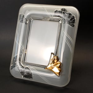 May include: A rectangular photo frame with a silver interior and a clear, frosted glass exterior. The frame features gold leaf accents and black leaf designs on the corners. White curved lines decorate the frame's surface.