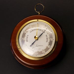 Könnte beinhalten: Ein rundes analoges Barometer mit einem polierten dunklen Holzrahmen und einer Messinglünette. Das weiße Zifferblatt zeigt schwarzen Text, darunter "REGEN", "VERÄNDERLICH" und "SCHÖN", sowie den Markennamen "Heckermann" und einen Metallring zum Aufhängen.