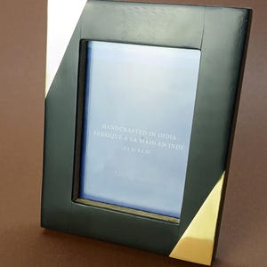 Peut inclure: Cadre photo noir rectangulaire avec des coins dorés et argentés. Le cadre a une finition sombre. Le texte "HANDCRAFTED IN INDIA, FABRIQUE À LA MAIN EN INDE" est visible, ainsi que la taille 3 x 4 / 8 x 10.