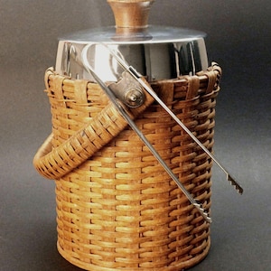 May include: A vintage ice bucket with a woven wicker basket design. It features a stainless steel lid and a wooden handle on top. The bucket includes a pair of stainless steel ice tongs. The bucket is approximately 8 inches tall.
