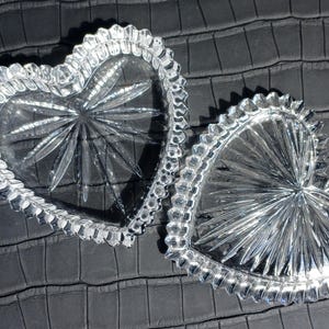 Pode incluir: Duas travessas de vidro transparente em forma de cora&ccedil;&atilde;o, com desenhos de estrela no centro e bordas onduladas. As travessas s&atilde;o exibidas em uma superf&iacute;cie texturizada cinza escuro, criando um contraste com o vidro brilhante.