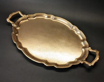 Puede incluir: Una bandeja de servicio ovalada de metal dorado con una superficie texturizada y un borde festoneado. La bandeja cuenta con dos asas decorativas en los extremos opuestos, cada una con un diseño curvo que combina con el borde festoneado.