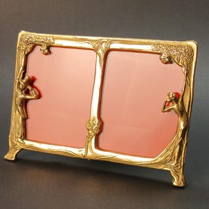 Könnte beinhalten: Ein doppelter Bilderrahmen mit goldener Oberfläche und rosa Hintergrund. Der Rahmen zeigt Figuren im Jugendstil und florale Details. Der Rahmen ist für zwei Fotos konzipiert und hat ein verziertes Design.