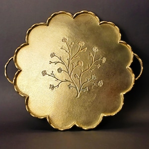 Peut inclure: Un plateau rond en métal doré avec un bord festonné et deux poignées incurvées sur les côtés opposés. Le centre du plateau présente un motif en relief représentant une plante ramifiée avec de petits boutons floraux sur une surface texturée.