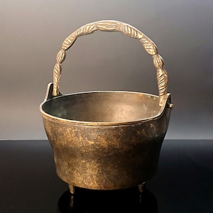 Peut inclure: Un chaudron en métal de couleur bronze avec une poignée incurvée et trois petits pieds. La poignée présente un motif de corde torsadée. Le bord du chaudron est mis en valeur par une garniture dorée. L'intérieur du chaudron est visible.