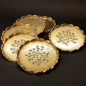 Pode incluir: Um conjunto de seis porta-copos circulares de metal dourado com bordas recortadas. Cada porta-copos apresenta uma superfície texturizada e um design central em relevo de uma árvore ramificada com pequenas flores, dispostos em grupo.