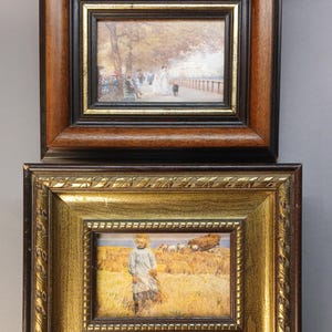 Op de afbeelding: Twee ingelijste landschapsschilderijen. Het bovenste frame is donker hout met een zwarte binnenrand, met een parkscène. Het onderste frame is goudkleurig met een gedetailleerde rand, met een kind in een veld. Beide zijn klein.