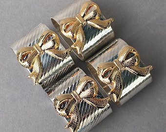 Könnte beinhalten: Vier silberne Serviettenringe mit strukturiertem Design und goldenen Schleifenakzenten. Die Serviettenringe sind quadratisch angeordnet und präsentieren ihr elegantes Design. Perfekt, um jedem Tisch eine elegante Note zu verleihen.