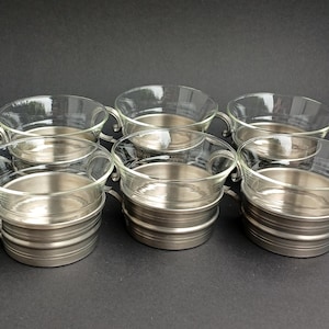 Puede incluir: Seis tazas plateadas con inserciones de vidrio. Las tazas tienen una forma redondeada y un acabado liso y brillante.