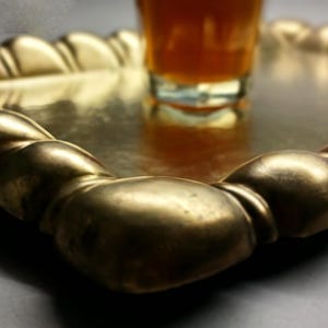 May include: A gold-colored, rectangular serving tray with a decorative, rounded edge. A glass with an amber liquid sits in the center of the tray. The tray is likely made of metal and has a reflective surface.