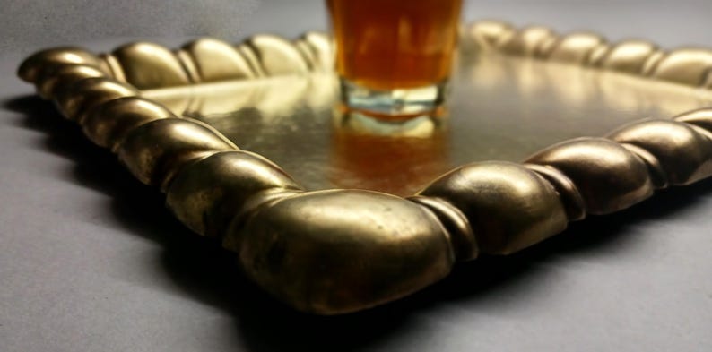 May include: A gold-colored, rectangular serving tray with a decorative, rounded edge. A glass with an amber liquid sits in the center of the tray. The tray is likely made of metal and has a reflective surface.