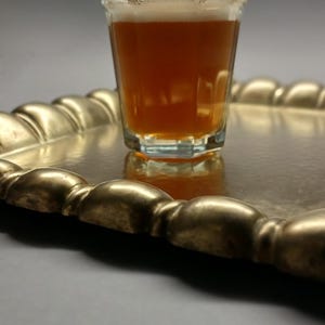 May include: A rectangular, gold-colored tray with a beaded edge holds a glass of amber liquid. The tray's surface reflects the glass and its contents. The tray is on a neutral background.