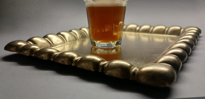 May include: A rectangular, gold-colored tray with a beaded edge holds a glass of amber liquid. The tray's surface reflects the glass and its contents. The tray is on a neutral background.