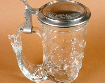 May include: Clear glass beer stein with a decorative metal lid and handle. The stein has a textured pattern on the glass and a hinged lid. The handle is shaped like a horn.