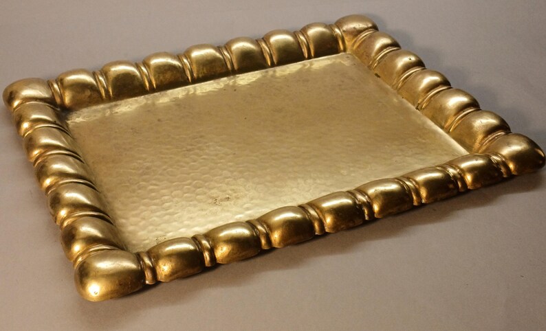 May include: A rectangular gold-colored tray with a hammered surface. The tray's border is adorned with a repeating, rounded design. The tray is likely made of metal and is suitable for serving or display.