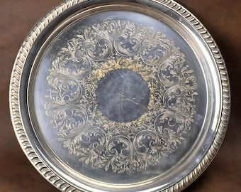 May include: A round silver serving tray with an ornate floral design in the center. The tray has a decorative rope-like edge. The interior of the tray has a dark, reflective surface.
