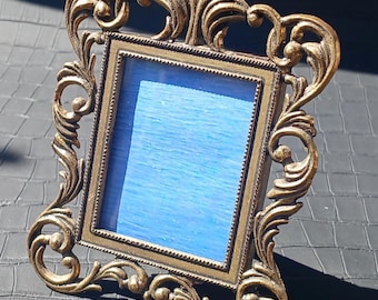 May include: Ornate, antique-style picture frame with a gold finish. The frame has a square opening with a blue background. The frame features intricate scrollwork and decorative flourishes, adding a vintage aesthetic.