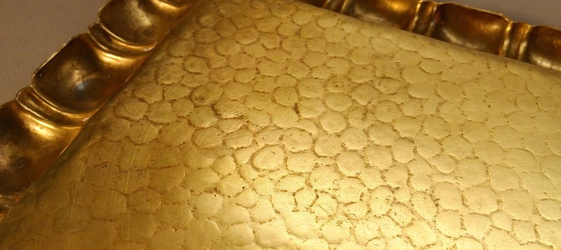 May include: Close-up of a gold-colored tray with a textured surface. The tray has a decorative, scalloped edge. The surface has a pattern of small, irregular circles.
