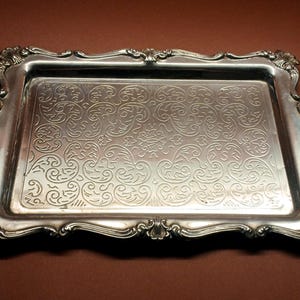 Può includere: Un vassoio rettangolare argentato con bordi ornati e arricciati e un motivo floreale in rilievo dettagliato sulla superficie. Il vassoio ha una finitura lucida e riflettente, su sfondo marrone.