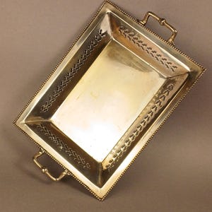 May include: A rectangular silver-coloured tray with two handles. The tray has a beaded border and decorative cut-out leaf patterns along the edges. The handles are a gold colour.