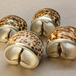 Może przedstawiać: Cztery ręcznie robione pierścienie do serwetek. Każdy pierścień ma naturalną muszlę z brązowym i beżowym wzorem w kropki, zamkniętą w srebrnej opasce. Otwarta konstrukcja ułatwia umieszczanie serwetek.