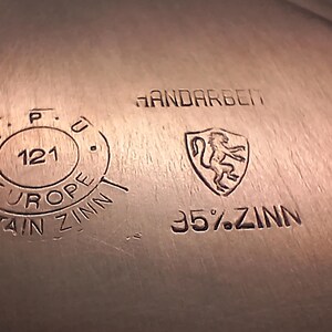 Puede incluir: Primer plano de una superficie met&aacute;lica con texto y s&iacute;mbolos estampados. El texto incluye "HANDARBEIT", "95%.ZINN" y "EUROPE ZINN". Tambi&eacute;n es visible un escudo con un le&oacute;n.