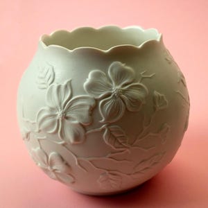 May include: A light-colored, round ceramic vase with a scalloped rim. The vase is adorned with embossed floral and leaf designs. The flowers are in full bloom, and the leaves have detailed veining. The vase is set against a pink background.