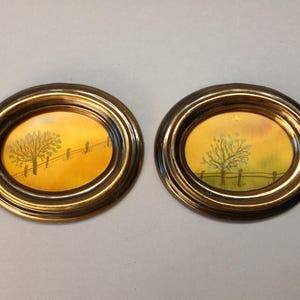 Op de afbeelding: Twee ovaalvormige wandlijsten met een metallic bronskleurige afwerking. Elke lijst bevat een klein landschapsschilderij met een boom, een hek en een warme gele en oranje lucht. Het kunstwerk toont een landelijk tafereel.
