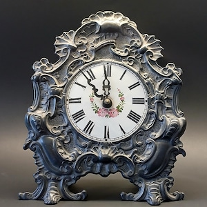 May include: A mantel clock featuring a dark grey, ornate metal frame with intricate scrollwork and relief detailing. The circular clock face is white with black Roman numerals, black hands, and a small floral wreath design in pink and green.