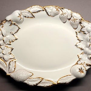 May include: An oval, white ceramic serving platter with a gold-trimmed border. The border is decorated with raised relief designs of grapes, strawberries, pears, and lemons. The platter is a decorative piece for serving food.