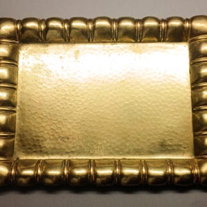 May include: A rectangular, gold-colored tray with a hammered surface. The tray's border is adorned with a series of rounded, segmented details. The overall design is symmetrical and ornate, suitable for decorative purposes.
