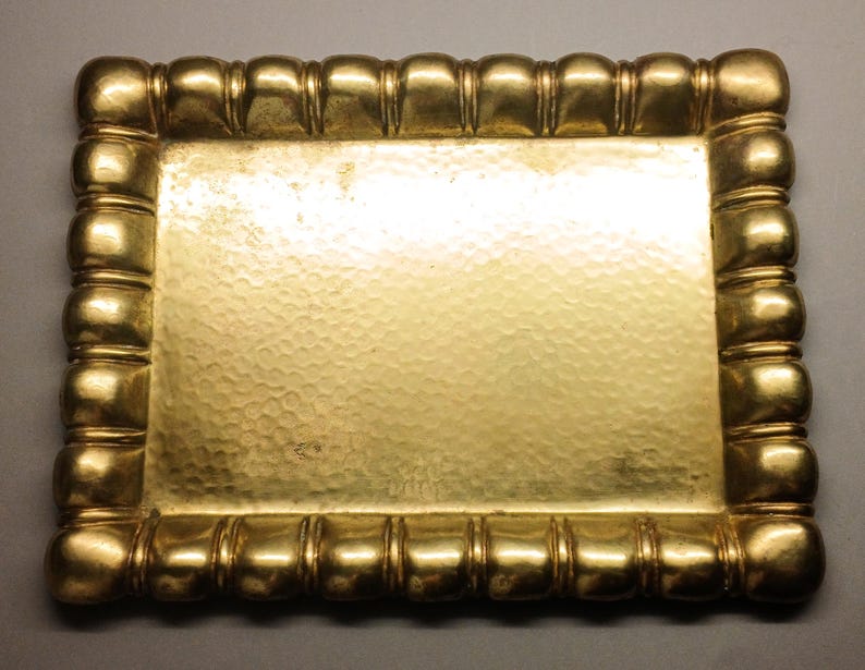 May include: A rectangular, gold-colored tray with a hammered surface. The tray's border is adorned with a series of rounded, segmented details. The overall design is symmetrical and ornate, suitable for decorative purposes.