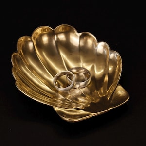 Könnte beinhalten: Eine polierte, goldfarbene Metallschale in Form einer Jakobsmuschel mit geriffeltem, geschwungenem Design. Zwei schlichte, silberfarbene Metallringe liegen in der Mitte der Muschelschale, die eine glatte, reflektierende Oberfläche aufweist.