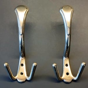 以下が含まれることがあります： 光沢のあるシルバーカラーの金属製ウォールフック2個。それぞれ中央に高い垂直のフックがあり、両側に2つの小さなフックが付いています。各フックのベースには取り付け用の円形のネジ穴が2つあり、全体的に滑らかで反射する仕上げが施されています。