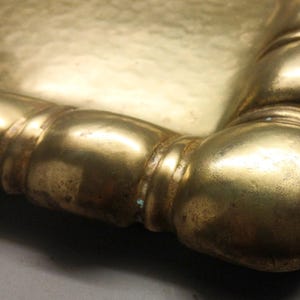 May include: Close-up of a gold-colored tray corner. The tray has a rectangular shape with rounded edges and a decorative, bulbous border. The metal has a slightly aged appearance.