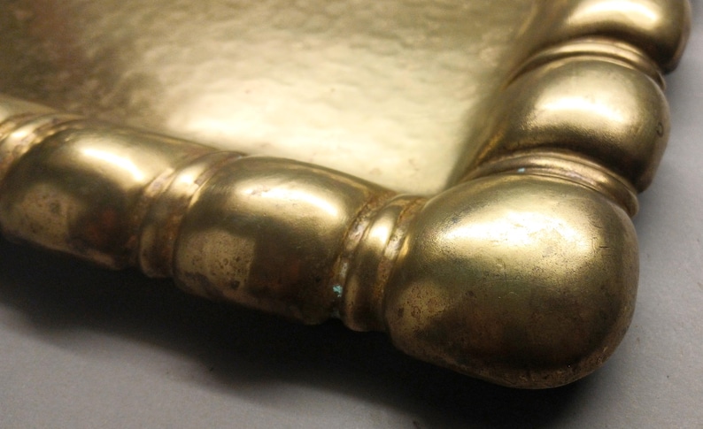 May include: Close-up of a gold-colored tray corner. The tray has a rectangular shape with rounded edges and a decorative, bulbous border. The metal has a slightly aged appearance.