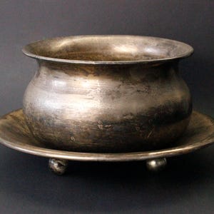 Pode incluir: Uma taça de metal prateado manchado com uma borda larga e alargada, apoiada numa base de prato circular a condizer. A taça apresenta um corpo arredondado e é suportada por três pequenos pés esféricos com um acabamento metálico desgastado.